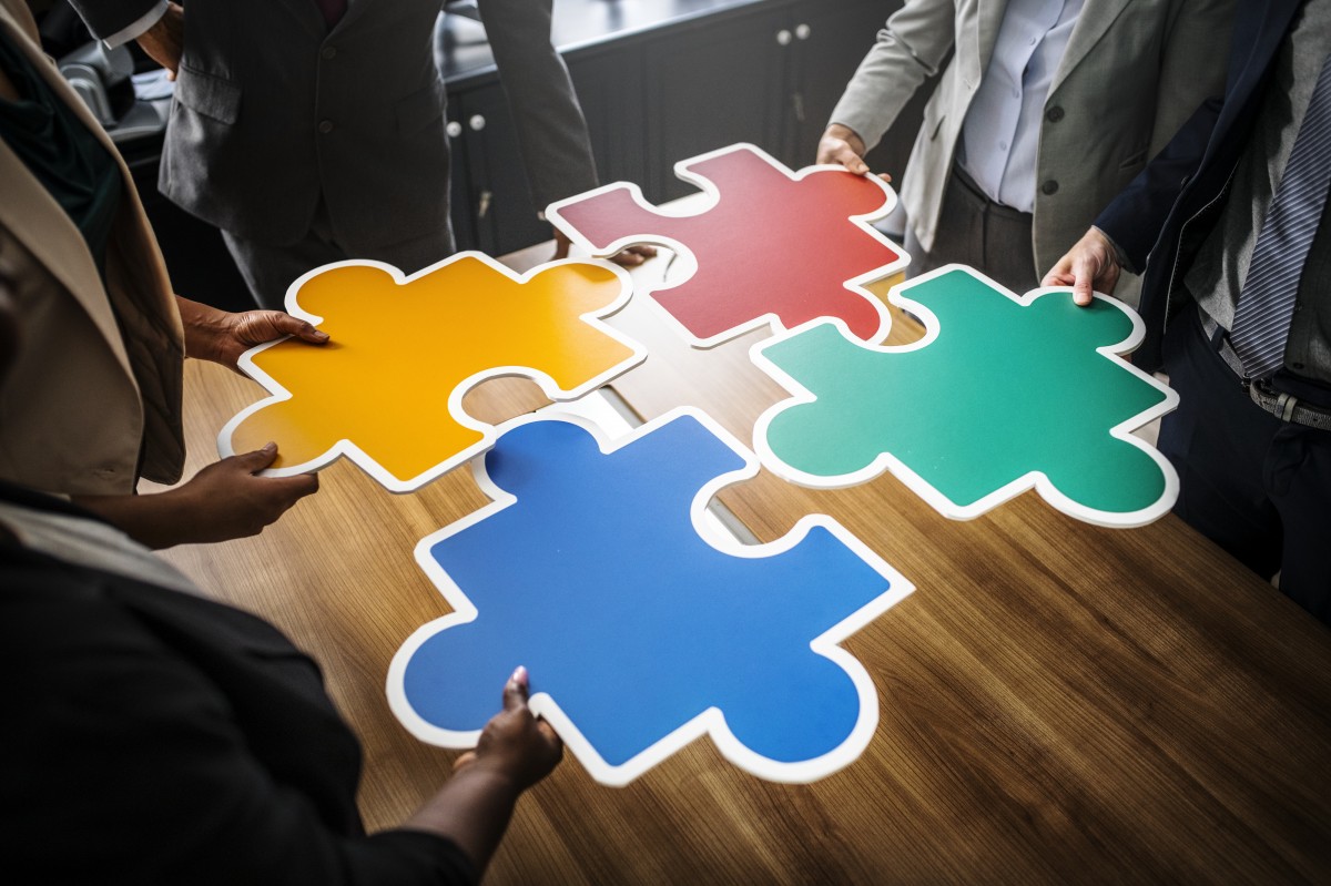 Four people with holding coloured puzzle pieces together over a table / Quatre personnes tenant ensemble des pièces de puzzle de couleur sur une table